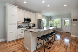 Uma cozinha moderna com armários brancos, uma ilha grande com banquetas, eletrodomésticos de aço inoxidável e um lustre.