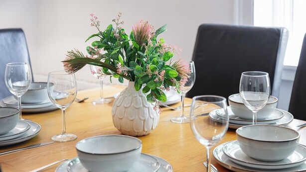 Una mesa de comedor dispuesta con platos blancos, cuencos y copas de vino transparentes, con un jarrón blanco en el centro adornado con follaje verde y flores rosas.