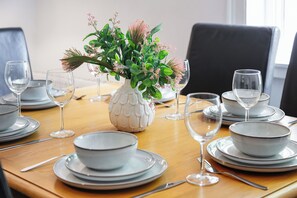 Ett matbord dukat med vita tallrikar, skålar och klara vinglas, med en vit vas i mitten fylld med gröna blad och rosa blommor.