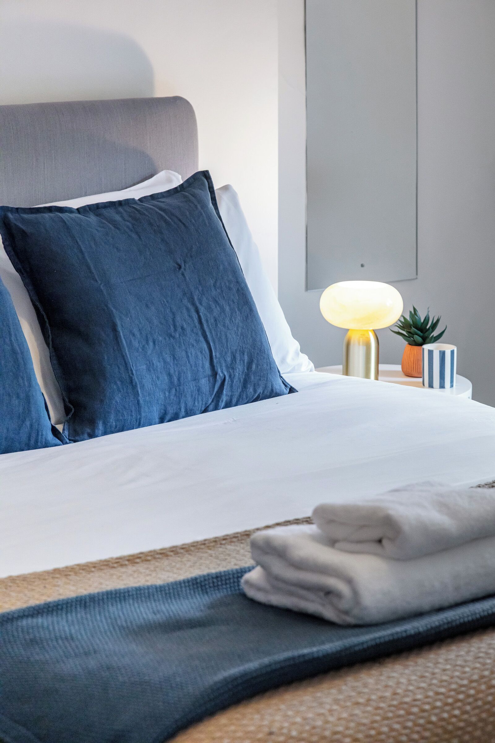 Un lit bien fait, avec une literie bleue et blanche, une lampe de chevet, une petite plante en pot et une tasse à rayures posée sur une table de chevet.
