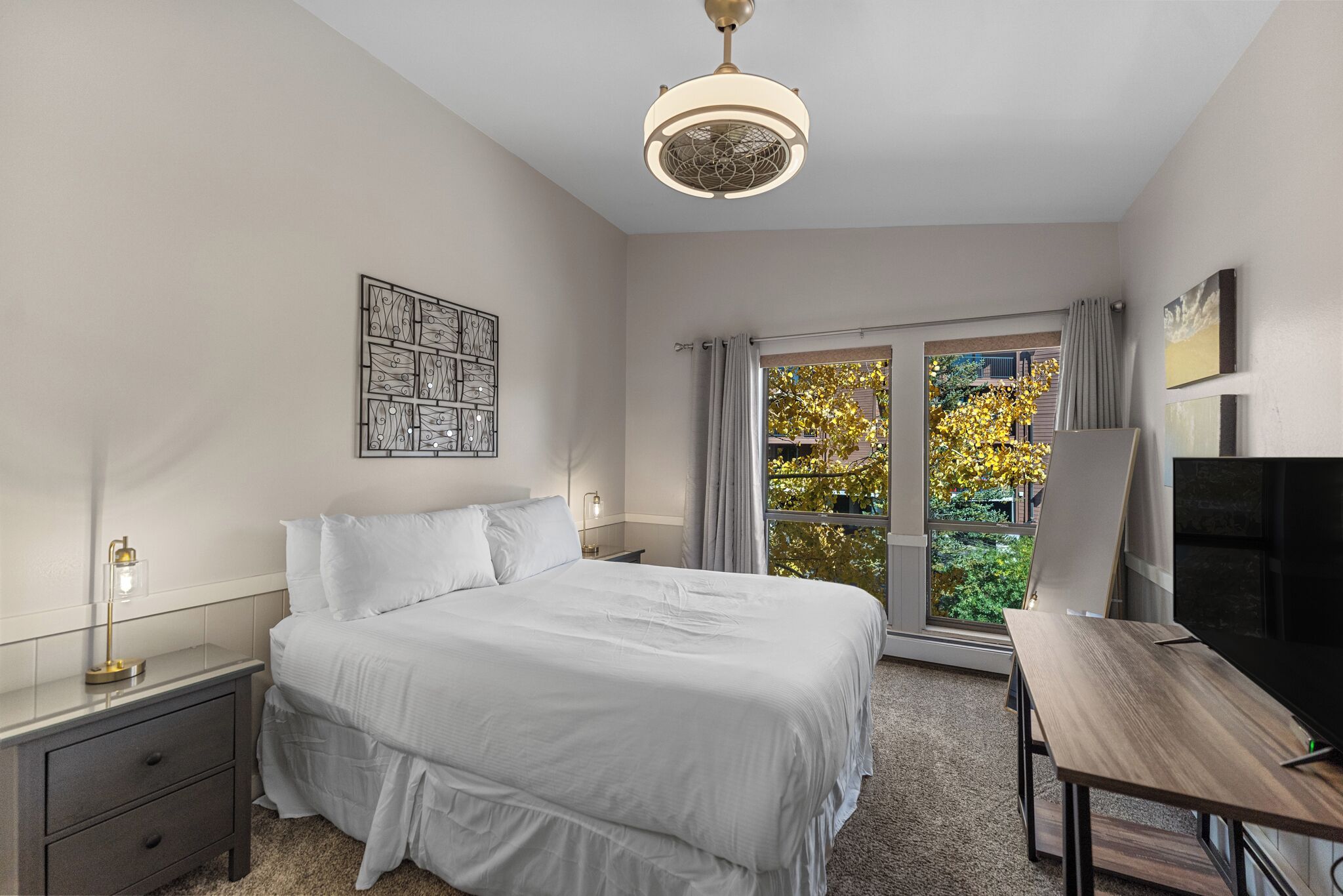 A bedroom with a bed, bedside table, lamp, and a view of the outdoors through a window.