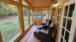 Een houten hut met grote ramen, een rieten bank en uitzicht op een bebost gebied.