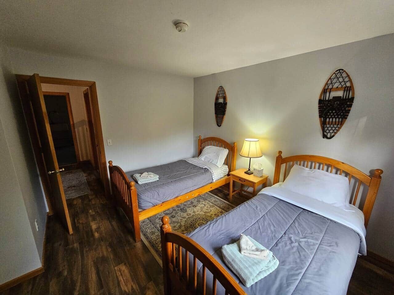 Een kamer met twee houten bedden, een nachtkastje met een lamp, en een deur die naar een andere kamer leidt.