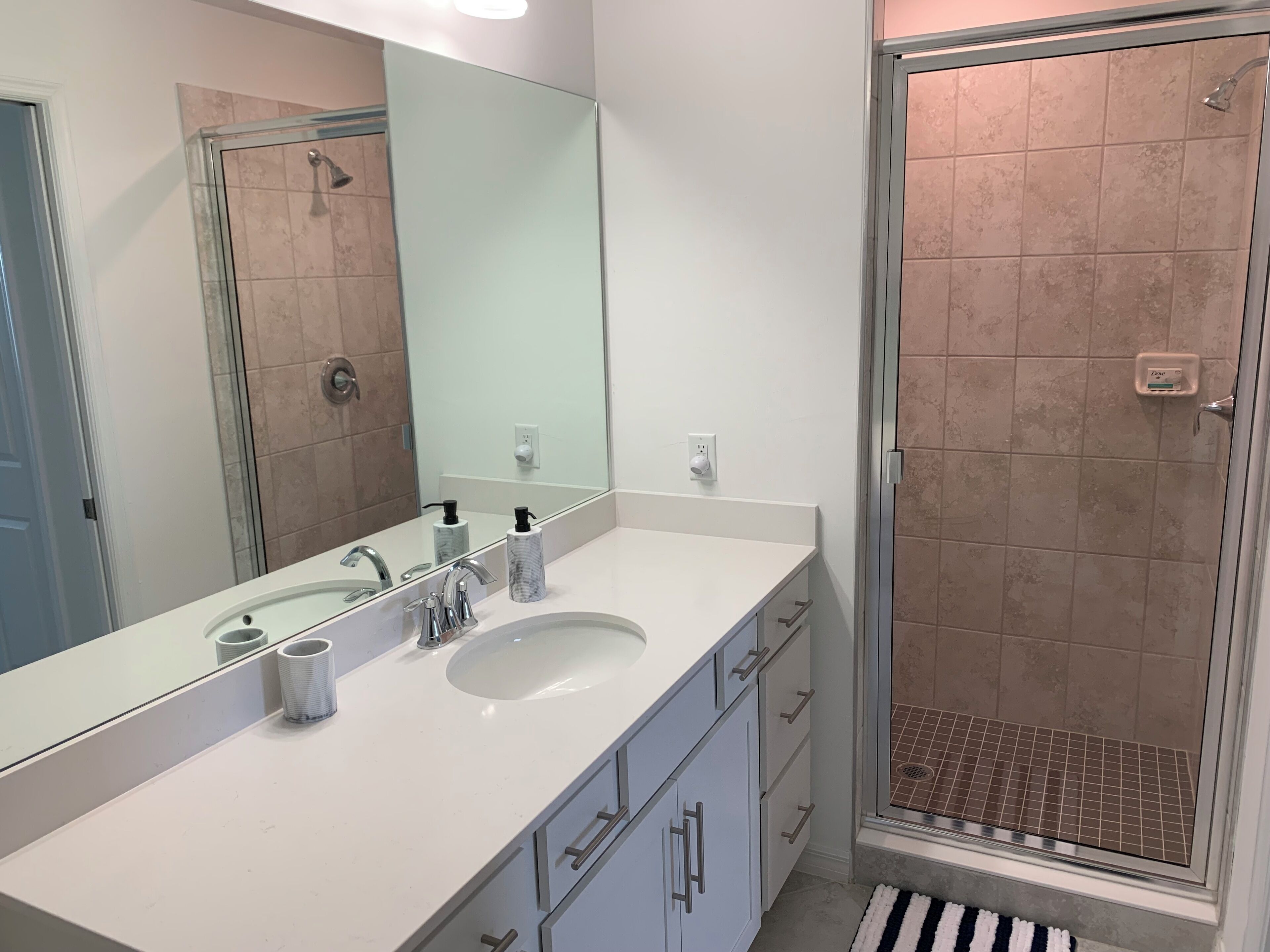 A modern bathroom with a white vanity, a large mirror, two sinks, and a glass-enclosed shower.