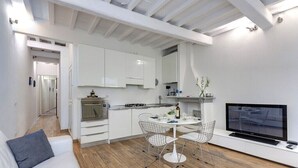 Una cucina moderna con mobili bianchi, un'area pranzo con tavolo rotondo e sedie in metallo, un divano con un cuscino e una TV a schermo piatto su un mobile bianco.