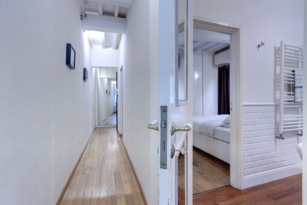 Een gang die leidt naar een slaapkamer met een bed, een radiator en een deur met een hendel.