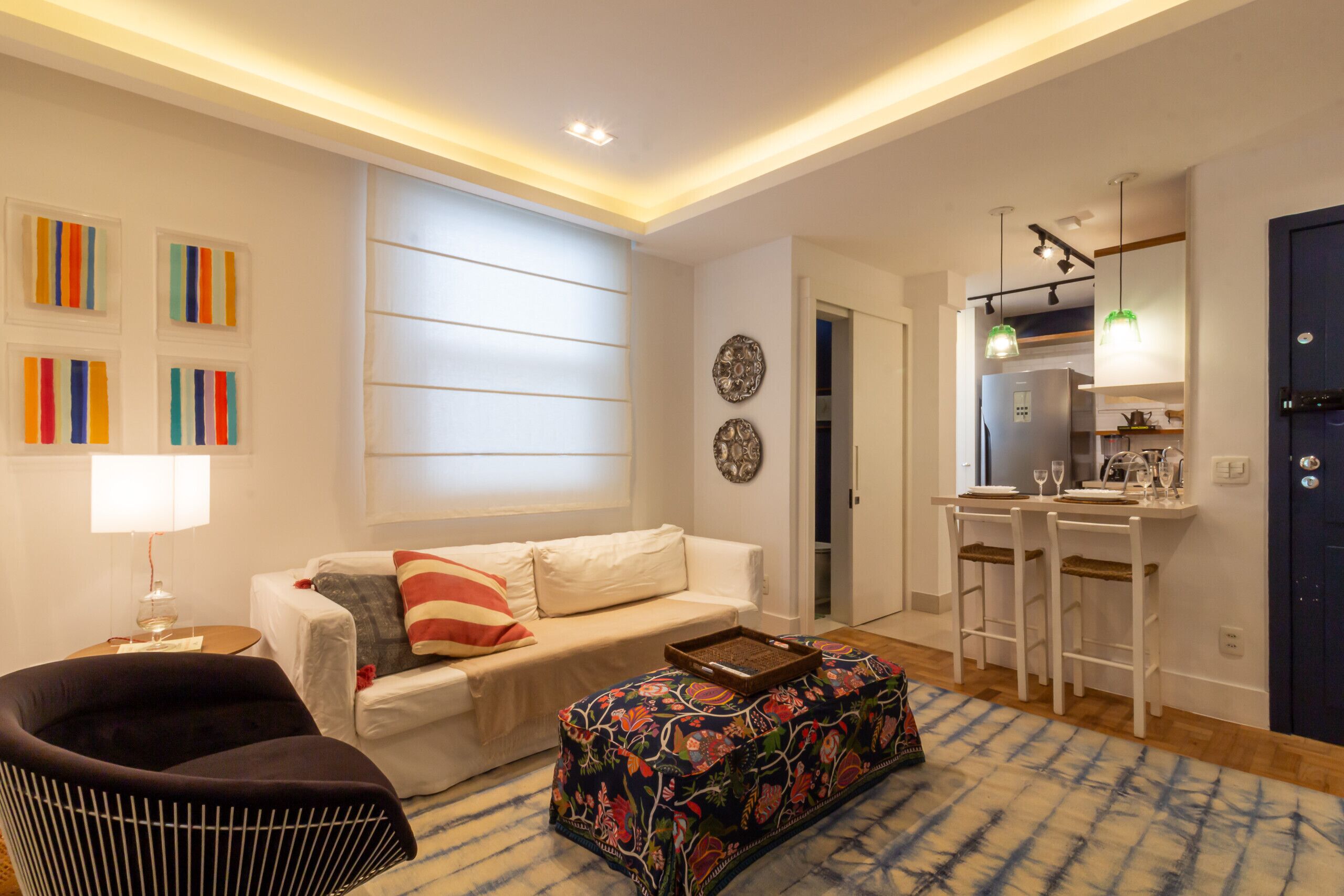 A modern living room with a sofa, a black chair, a patterned ottoman, and a dining area with bar stools.