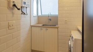 Eine kleine Küche mit weißen Fliesen, einem Fenster, einem Waschbecken, einer Waschmaschine und einer Toilette.