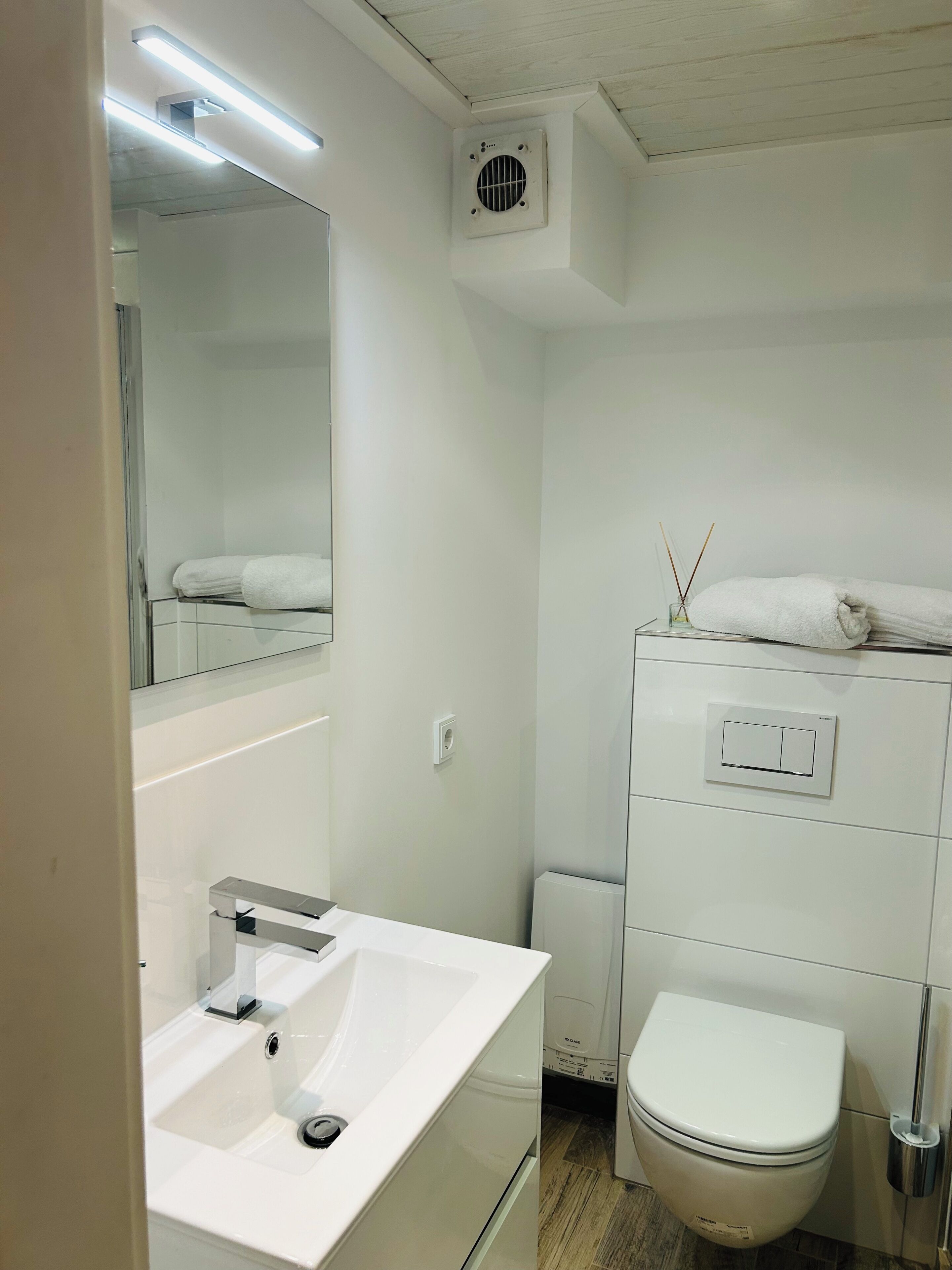 A modern bathroom with a white sink, mirror, toilet, and a small cabinet with a towel.