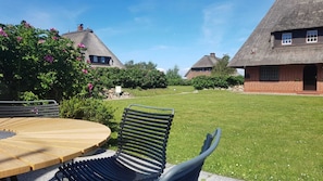 A well-maintained lawn with a wooden table and metal chairs, surrounded by thatched-roof houses and lush greenery.