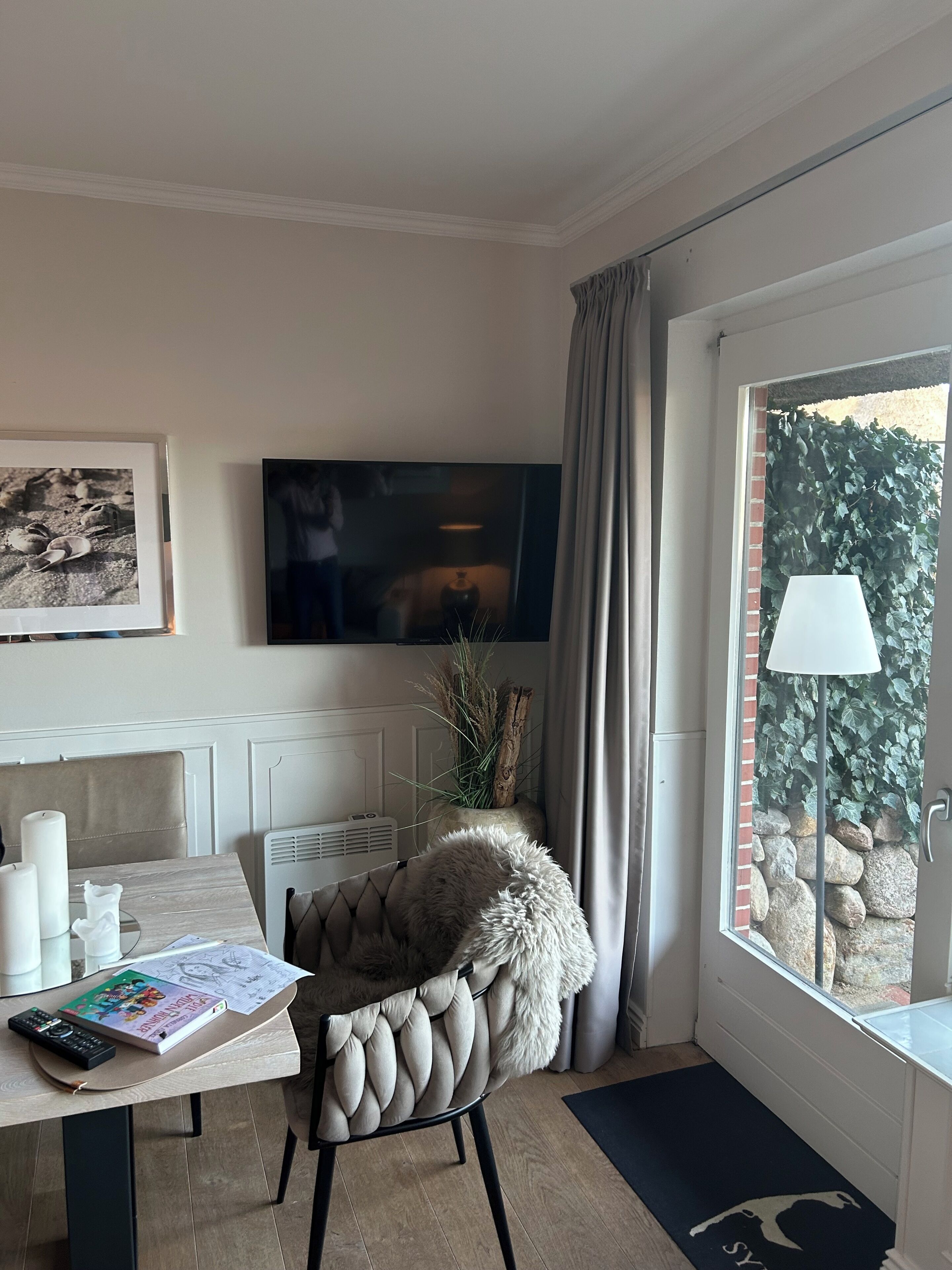 A room with a TV, a chair with a fur throw, a table with a book and remote, and a window with a view of greenery.