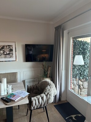 Una habitación con televisor, una silla con una manta de piel sintética, una mesa con un libro y un control remoto, y una ventana con vistas a la vegetación.