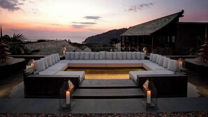 Zona relax esterna moderna con divano e cuscini bianchi, circondata da lanterne, con vista su un paesaggio costiero al tramonto.