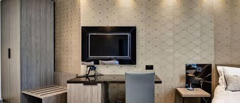 Habitación compacta con escritorio integrado, televisor montado en la pared, cama con cabecera azul y un papel tapiz estampado.