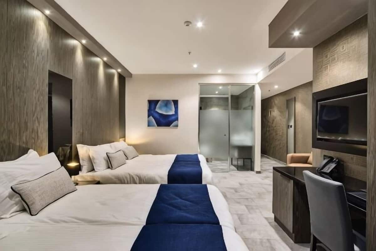 A hotel room with two beds, a TV, a desk, and a glass-enclosed shower area.