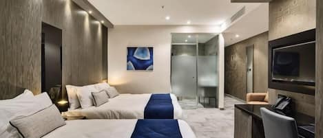 A hotel room with two beds, a TV, a desk, and a glass-enclosed shower area.