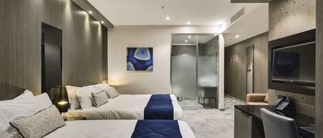 Habitación de hotel con dos camas, televisor, escritorio y baño con puerta de vidrio.