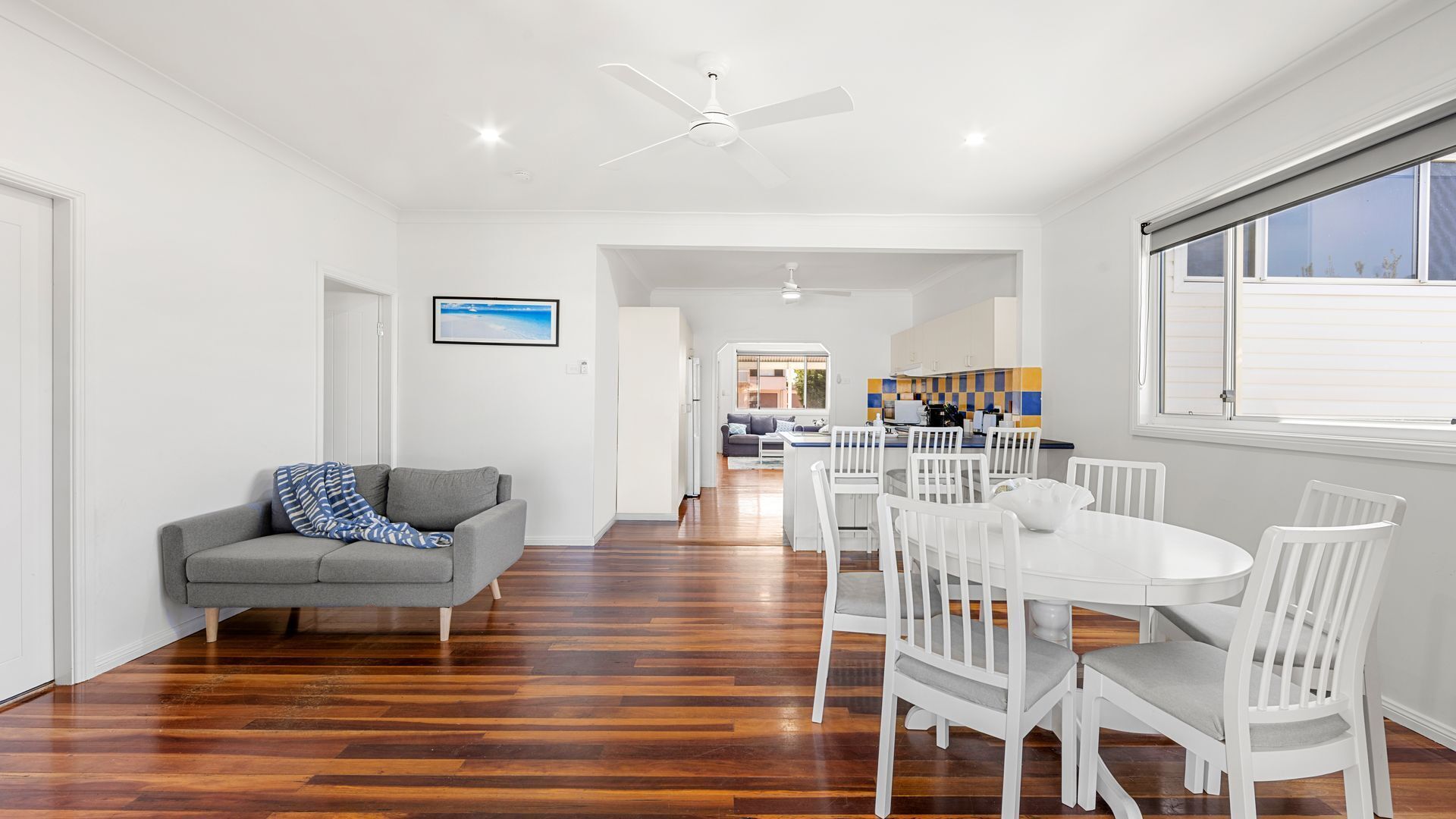 A modern living room with a grey sofa, white dining set, wooden flooring, and a ceiling fan.