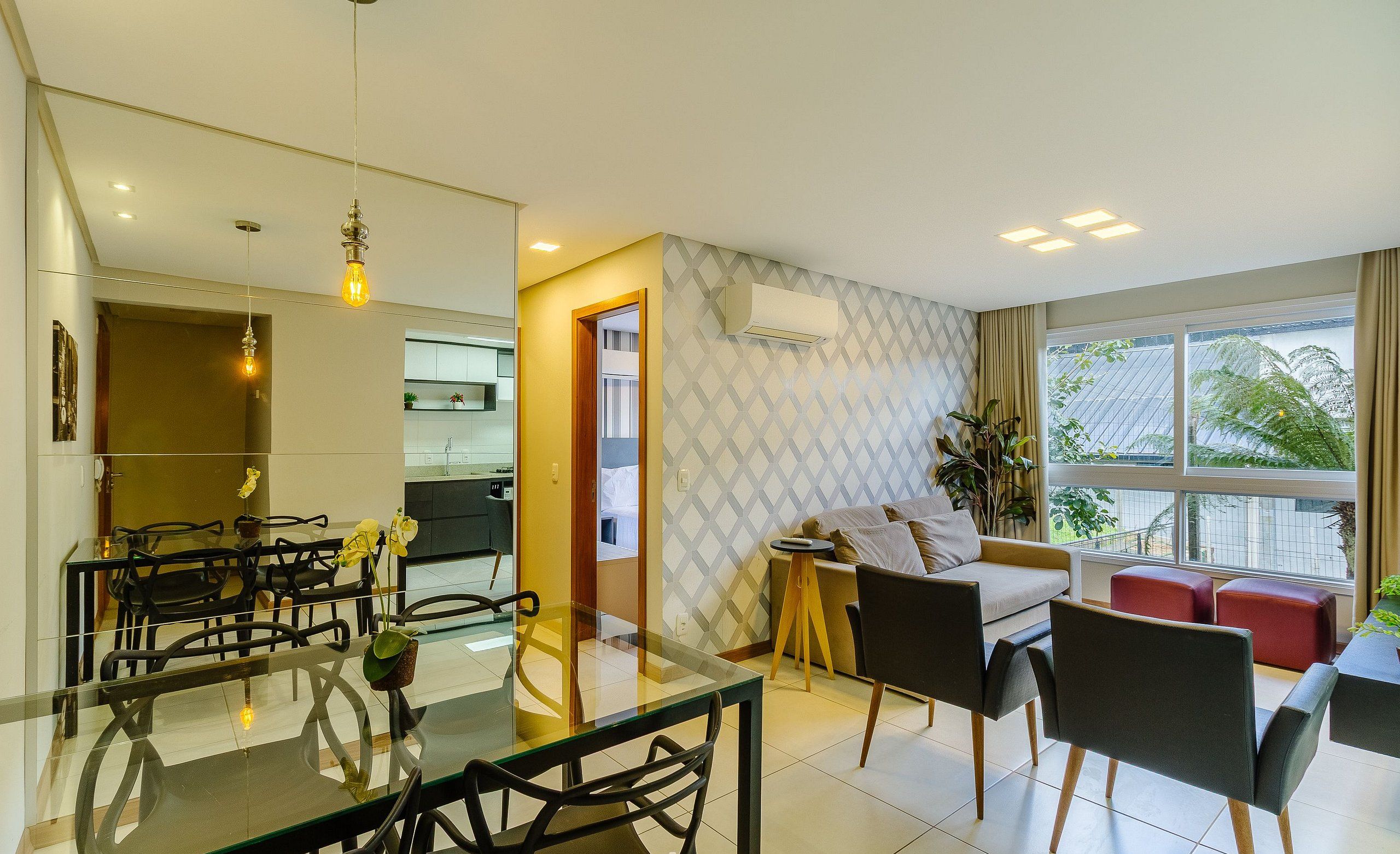 Una sala moderna con una mesa de comedor de vidrio, sillas negras, un sofá y un reposapiés rojo. Se observa una cocina al fondo y un amplio ventanal con cortinas.