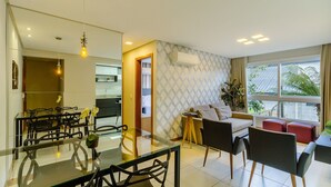 Ein modernes Wohnzimmer mit einem Glastisch, schwarzen Stühlen, einer Couch und einem roten Hocker. Im Hintergrund ist eine Küche erkennbar und ein großes Fenster mit Vorhängen.