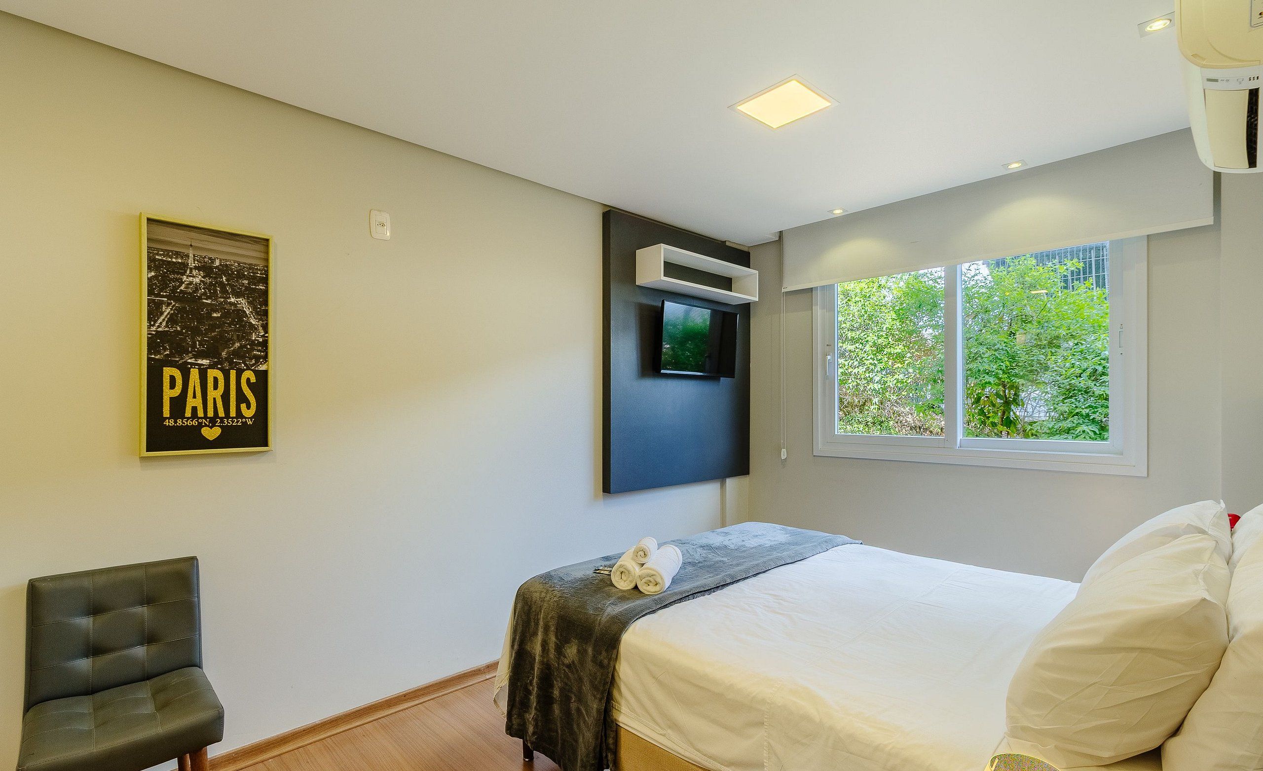 A modern bedroom with a bed, a chair, a TV mounted on the wall, and a window with a view of trees.