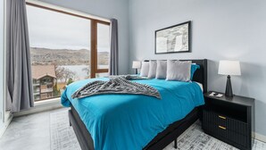 A bedroom with a large window, a bed with blue bedding, a nightstand with a lamp, and a framed picture on the wall.