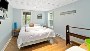 A bedroom with a bed, a TV mounted on the wall, two windows, a bedside table, and a nightstand. - Charming Cottage on the Lake - Family and Pet Friendly (LaBelle)