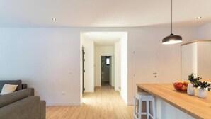 A modern living room with a grey sofa, a wooden floor, a kitchen area with a wooden countertop and white stools, and a hallway leading to another room.