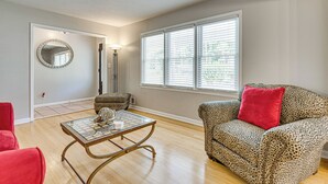 A living room with a glass coffee table, a leopard print sofa with a red pillow, a red chair, a leopard print ottoman, and a round mirror on the wall. - 3 Mi to Arabia Mtn Trails: Quiet Ranch-Style Home (Stonecrest)