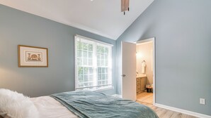 Ein Schlafzimmer mit Bett, einem Fenster mit Jalousien, einem gerahmten Bild an der Wand und einer Tür zu einem Badezimmer.