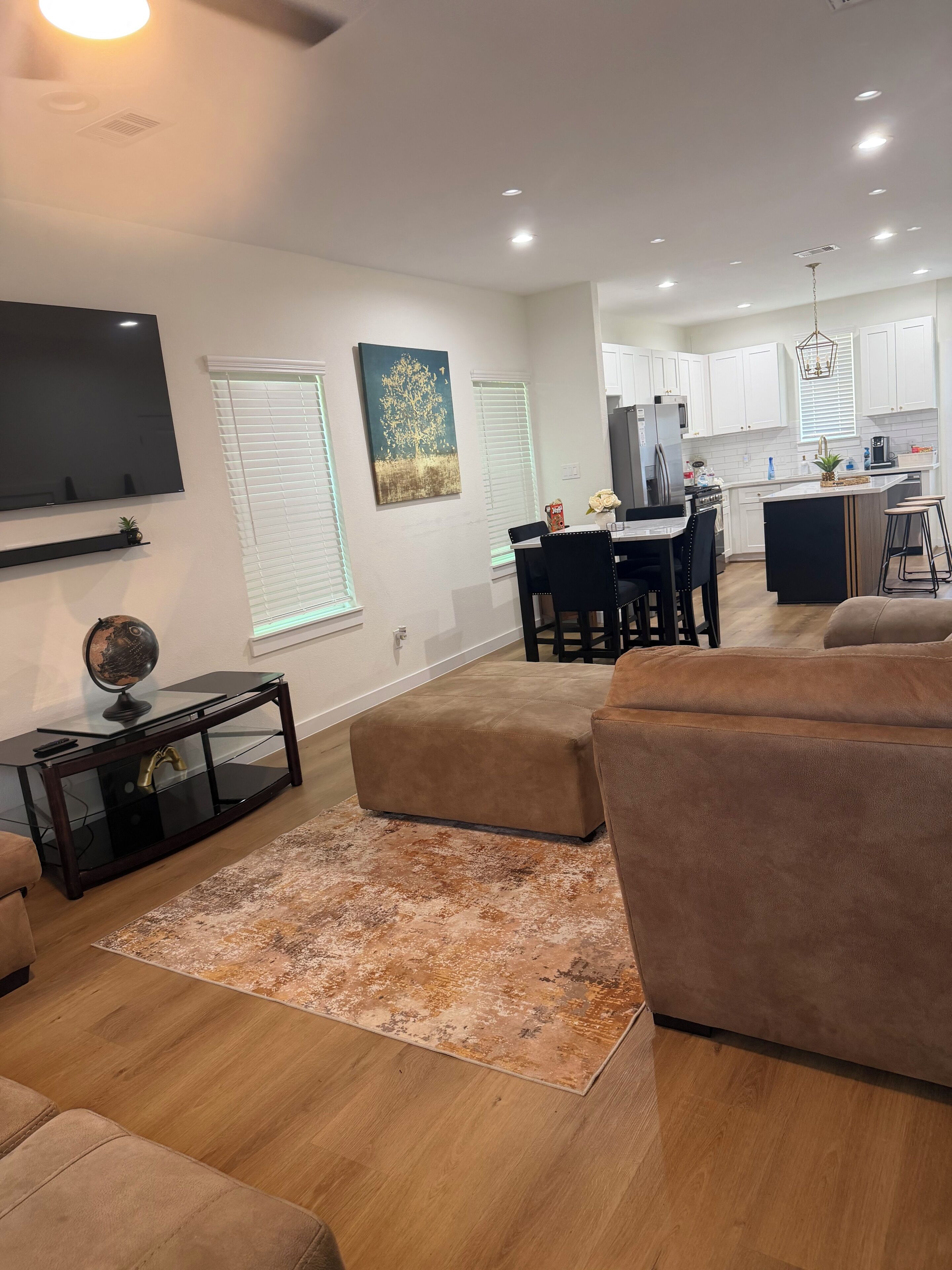 A modern living room with a flat-screen TV, a coffee table, and a sofa. There is a dining area with a table and chairs, and a kitchen in the background.