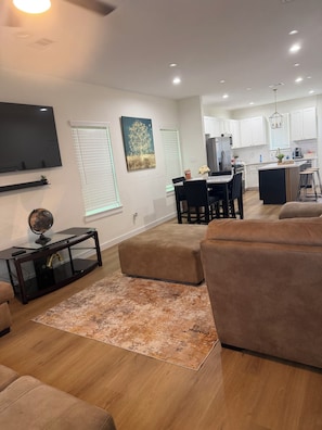 A modern living room with a flat-screen TV, a coffee table, and a sofa. There is a dining area with a table and chairs, and a kitchen in the background. - Houston Haven near all you need /A/ (Houston)