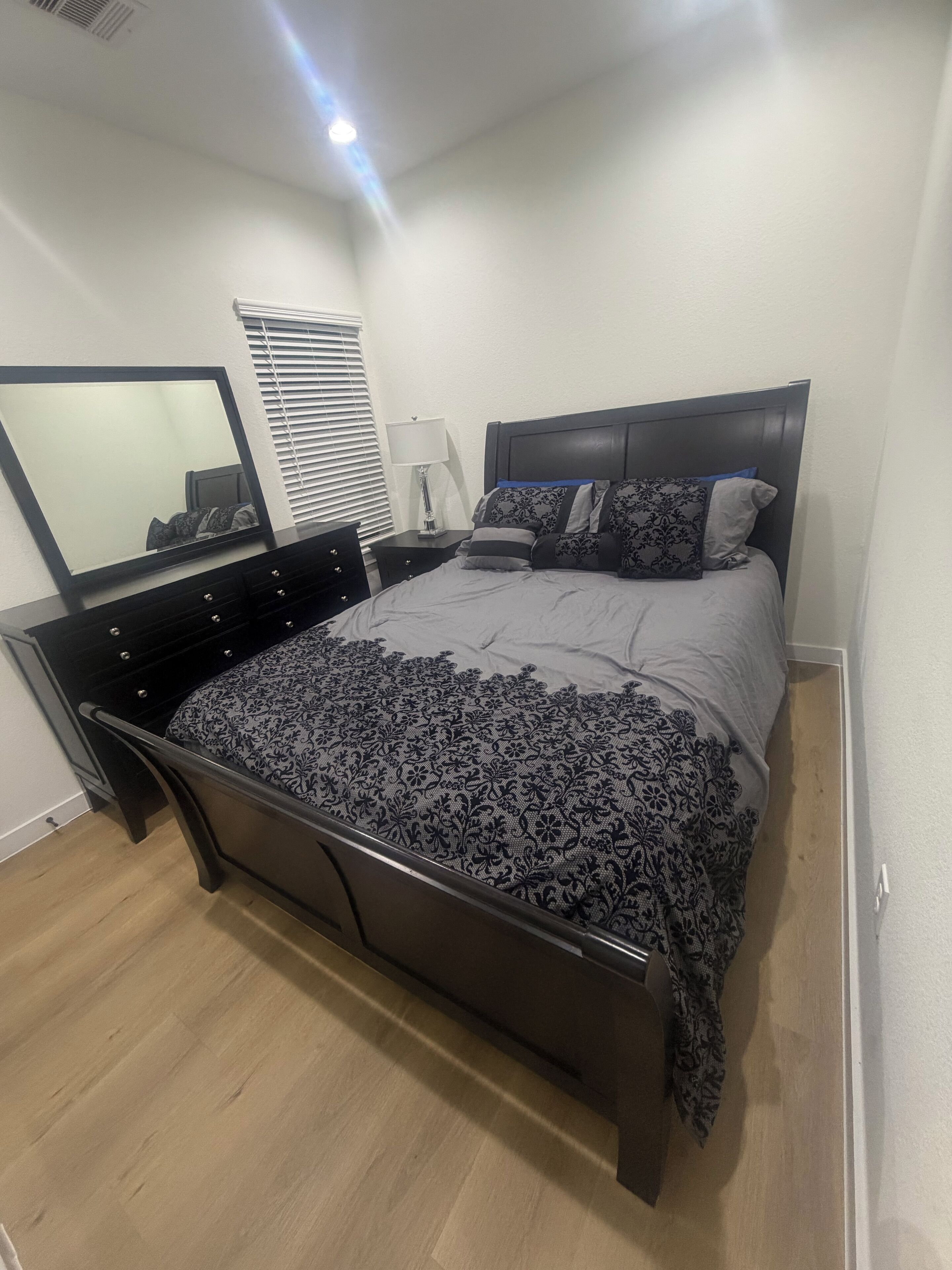 A modern bedroom with a large bed, a nightstand, a mirror, and a window with blinds.