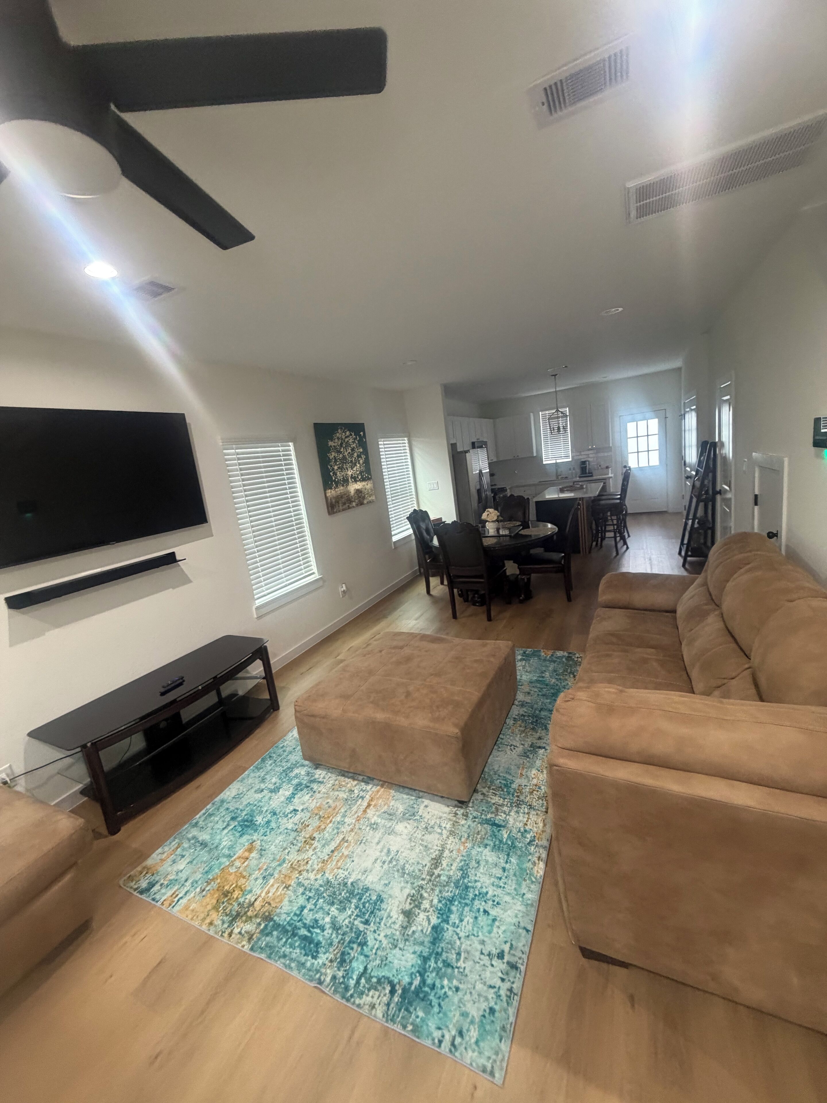 A spacious living room with a large flat-screen TV, a brown sectional sofa, and a coffee table. There is a dining area with a table and chairs in the background.