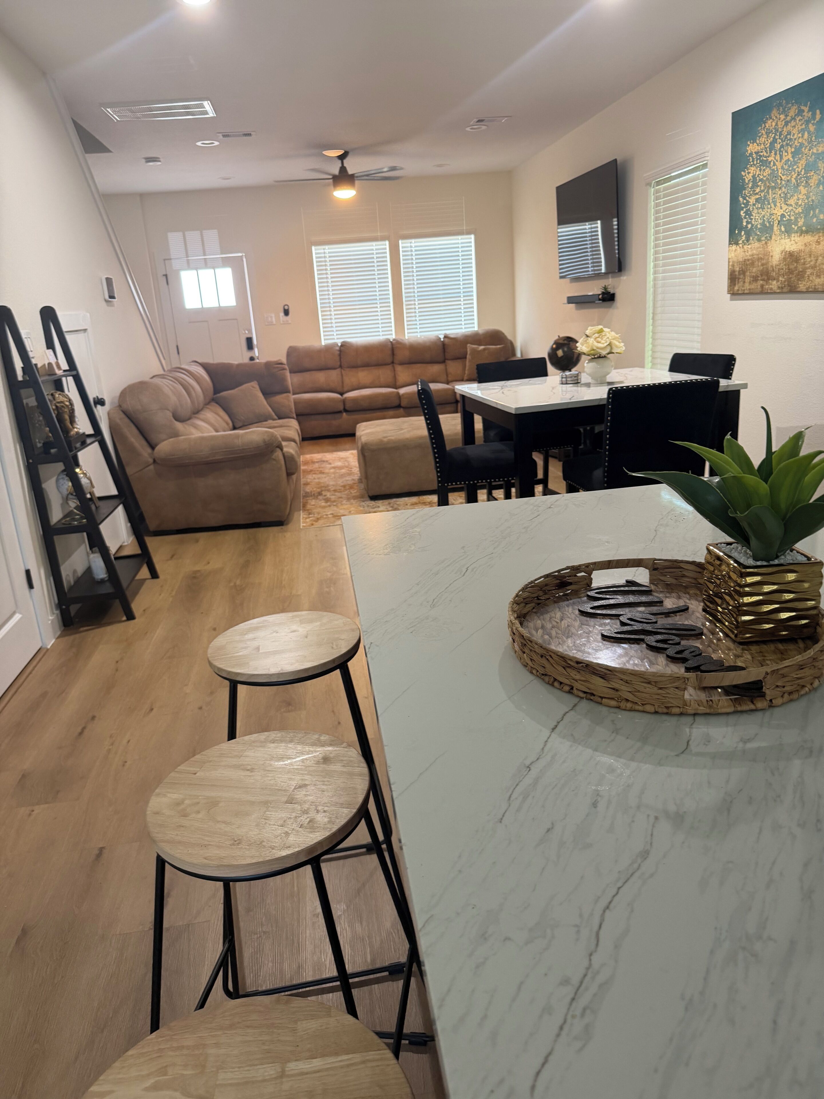 A modern kitchen with a marble countertop, two stools, a dining table with chairs, and a ceiling fan.