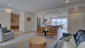 Una sala de estar moderna con una mesa de centro redonda de madera, un sofá gris y dos sillones. Se ve una cocina en el fondo, con alacenas blancas y electrodomésticos de acero inoxidable.
