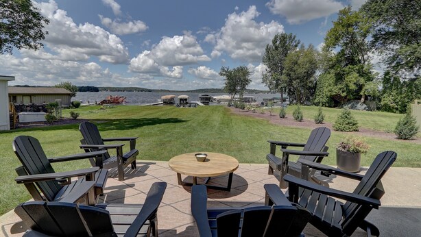 Un patio con sillas Adirondack y una mesa redonda, con vista a un lago con barcos y una zona verde.