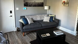 A modern living room with a grey sofa, patterned cushions, a black coffee table with books, a cityscape painting, a floor lamp, and a door with a light switch. - Mid-Century Tudor in Quiet Detroit Neighborhood (Detroit)