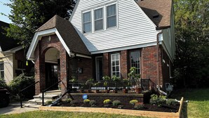 A two-story house with a brick facade and white siding, featuring a front porch with potted plants and a small garden. - Mid-Century Tudor in Quiet Detroit Neighborhood (Detroit)