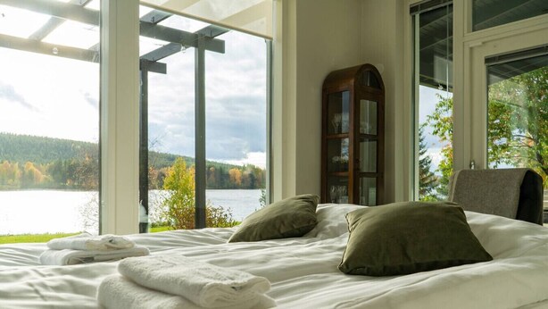 Een slaapkamer met een groot raam met uitzicht op een meer en bos, een bed met witte lakens en een houten kast.