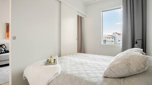 Une chambre d’hôtel avec un grand lit, une fenêtre donnant sur des immeubles, une table de chevet avec un plateau, et une chaise avec un coussin.