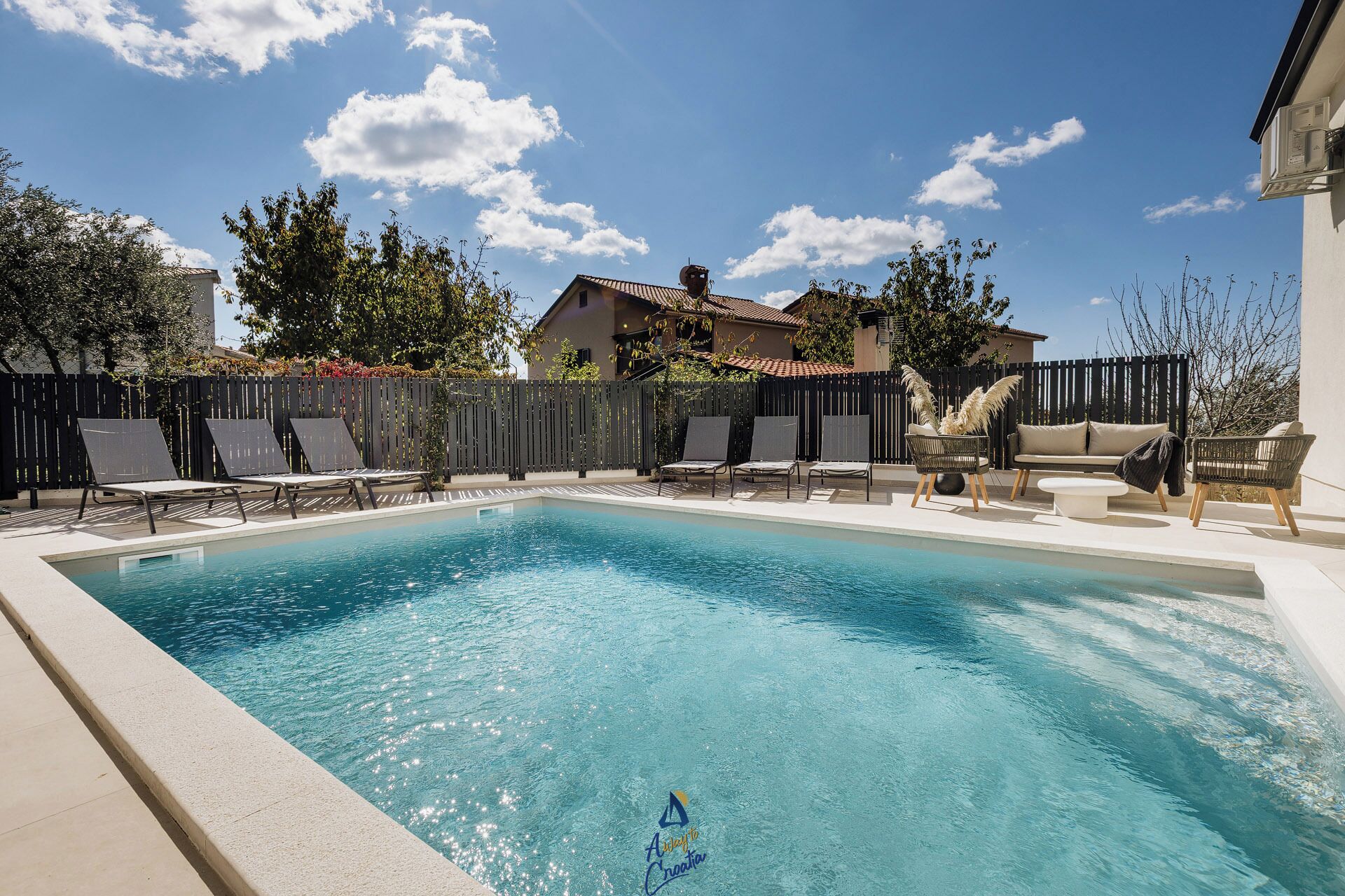 A rectangular swimming pool with a tiled edge, surrounded by lounge chairs and a sofa, with a fence and trees in the background.