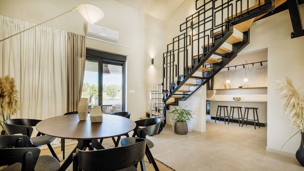 A modern interior with a round dining table, black chairs, a staircase with a metal railing, and a kitchen area with stools. - Charming New 3BR Villa for 6+2 with Private Pool (Višnjan)
