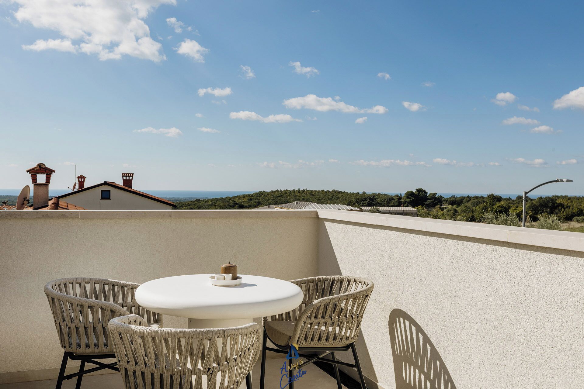 A terrace with a round table and wicker chairs, overlooking a coastal landscape with buildings and vegetation.