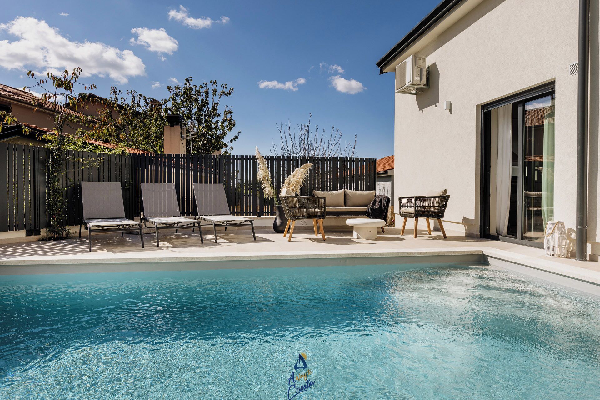 Une piscine entourée d’une clôture et de verdure, avec des chaises longues et un canapé à proximité.