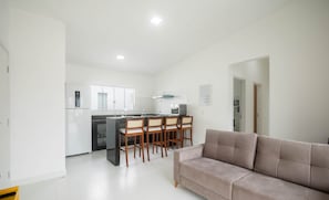 A modern living room with a beige sofa and a kitchen area featuring a dining table with chairs. - Fully Equipped Apartments in a Condo With a Pool, 750m From the Beach - Vifs (Porto Seguro)