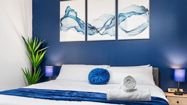 A neatly made bed with white linens, a blue throw blanket, and a blue pillow. There are three wall-mounted paintings above the bed, a potted plant to the left, and a nightstand with a lamp to the right. - Cozy King Suite|Central, Workspace + Free Parking (Pittsburgh)