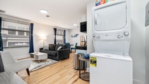 A modern living room with a grey sofa, a white coffee table, and a black armchair. There is a white washing machine in the corner and a small trash bin next to it. - Cozy King Suite|Central, Workspace + Free Parking (Pittsburgh)