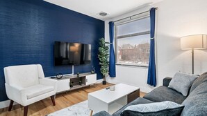 A modern living room with a blue accent wall, a flat-screen TV, a white armchair, a grey sofa, a coffee table, and a potted plant. - Cozy King Suite|Central, Workspace + Free Parking (Pittsburgh)