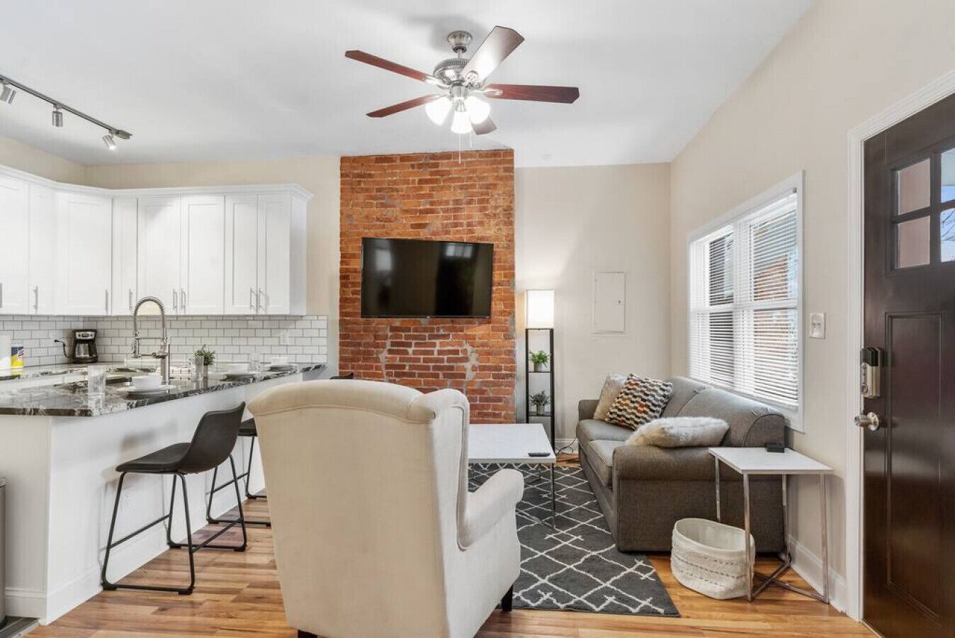 A modern kitchen with white cabinets, a brick accent wall, a ceiling fan, and a flat-screen TV mounted above the counter.